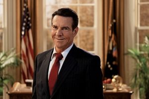 Dennis Quaid as Ronald Reagan in his film "Reagan", standing in his Oval Office in black tux and red tie and smiling at the camera.