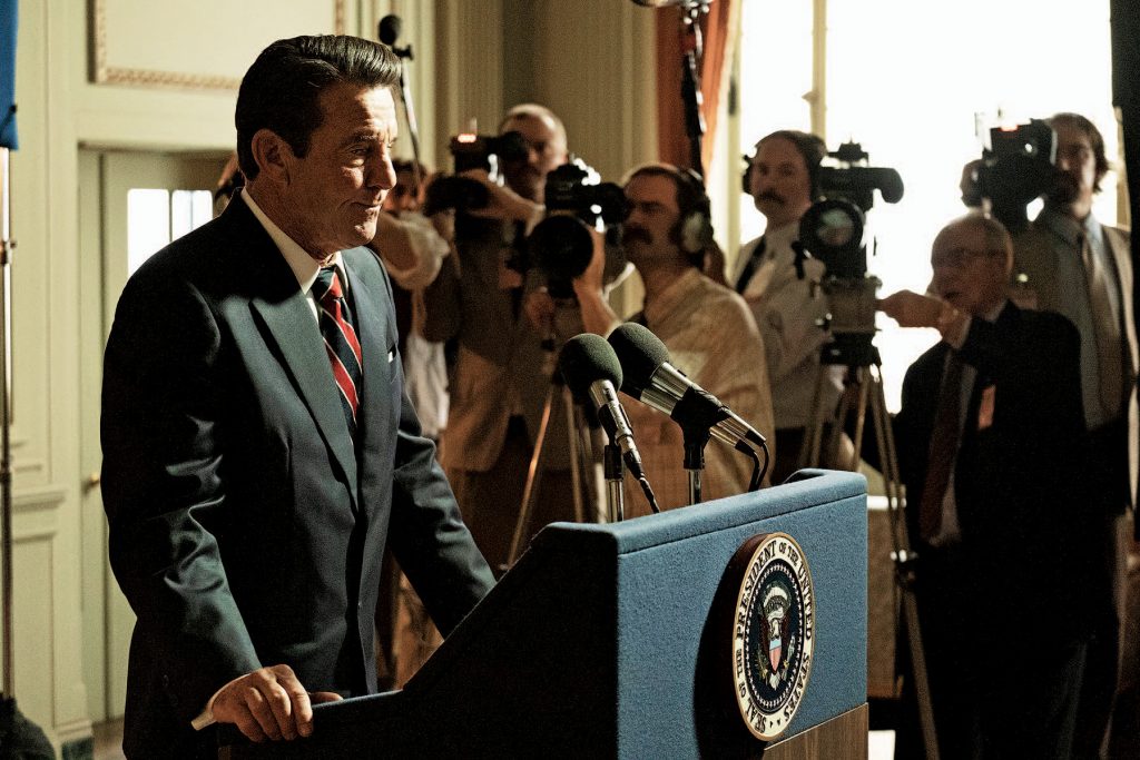 Dennis Quaid as Ronald Reagan in his film "Reagan", standing behind a podium in front of a press conference, with cameras surrounding.