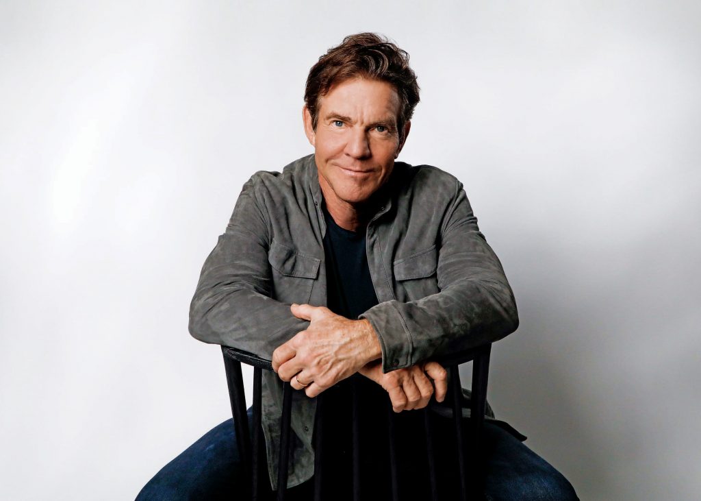 Dennis Quaid sitting in a chair and smiling to the camera. He wears jeans, a black shirt, and a gray jacket.