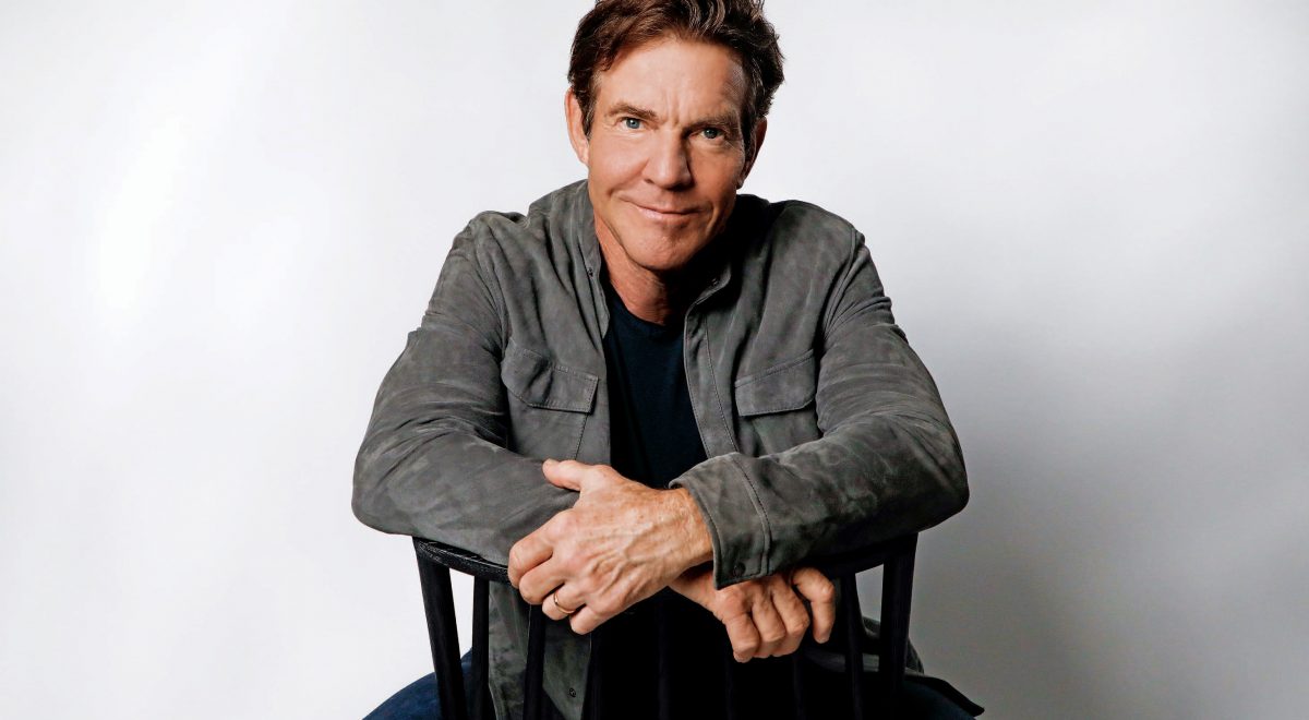 Dennis Quaid sitting in a chair and smiling to the camera. He wears jeans, a black shirt, and a gray jacket.