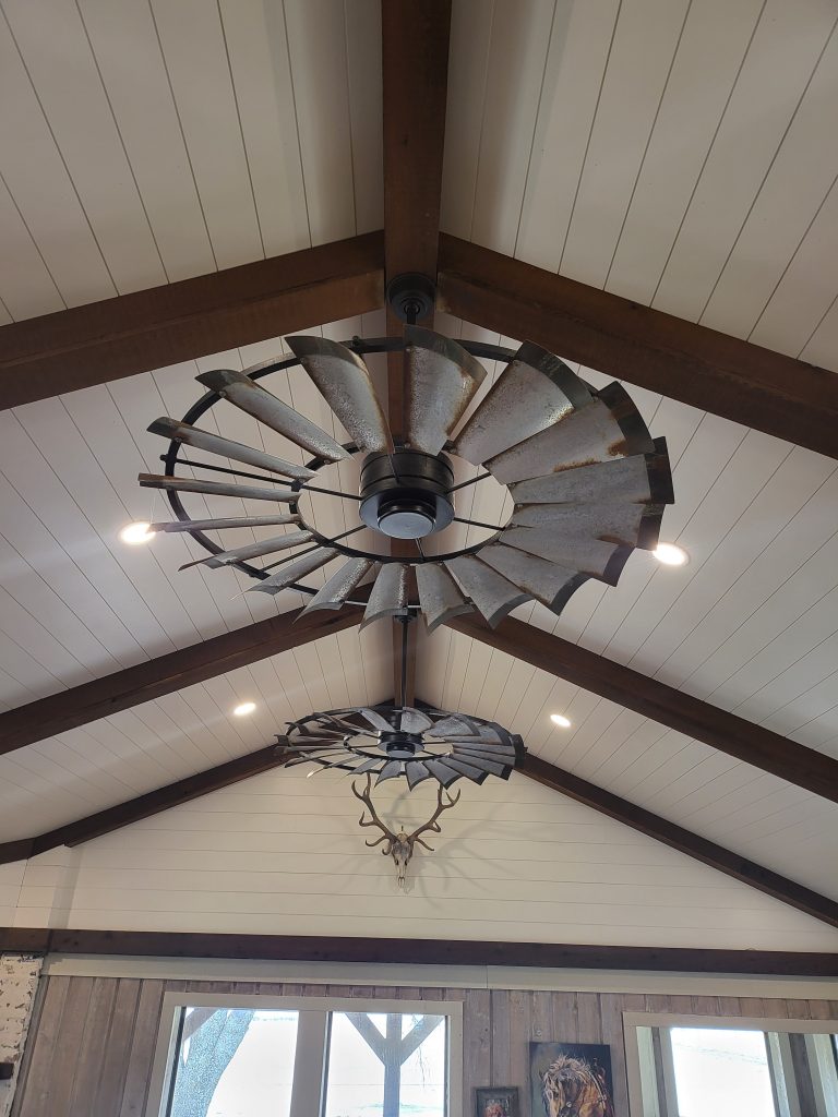 A Cattleman Windmill Ceiling Fan hanging from a white ceiling. It is silver with wood colored edging.