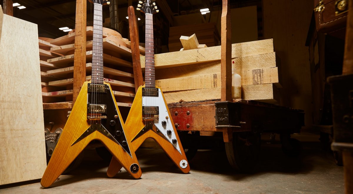 Two Gibson Custom Shop 1958 Korina Flying V Reissues (one in Black and White) behind the backdrop of a Gibson warehouse.