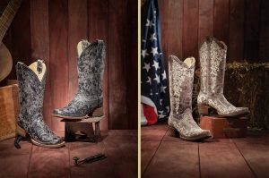 A split screen of a pair of black luxury Corral boots with white embroidery (left) and a pair of brown luxury Corral boots with white embroidery (right), both against a barn wall background.