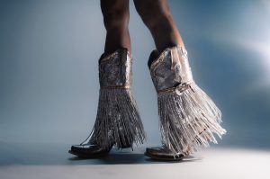 A pair of silver Corral boots against a silver background, with fringe hanging from midway down the boot.
