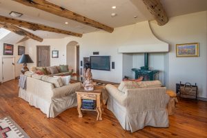 The secondary gathering space of the Kokopelli Ranch house, featuring two large couches and a lounge chair centered around a furnace, with rustic finishes.