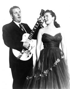 Les Paul playing a guitar and singing with Mary Ford at his side singing along with him circa 1954.