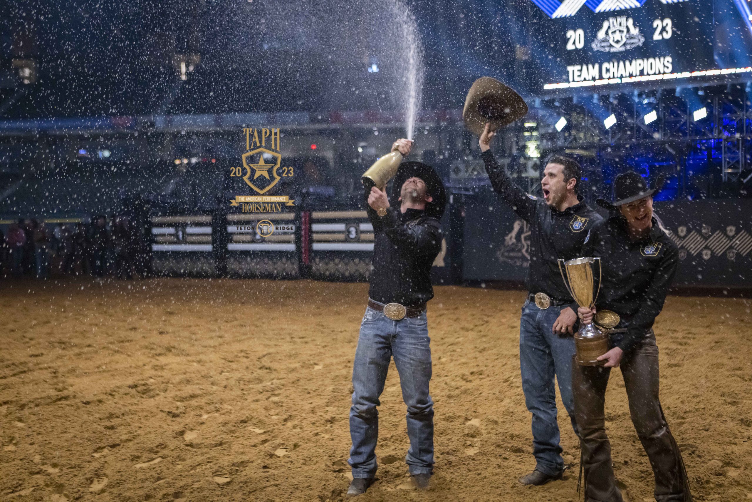 Adan Banuelos (left) sprays champaign in celebration with Fernando Salgado (middle), who holds his hat up in celebration, and Sarah Dawson (right), who holds the American Performance Horseman trophy.