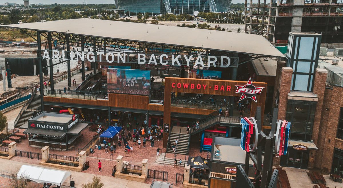 A large two-story open-air venue with a large sign above it saying Arlington Backyard. Two large TVs can be seen along with a bar area called Balcones and a smaller sign reading Cowboy Bar above the second floor.