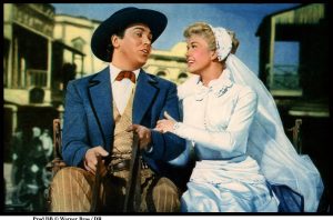 Actors Doris Day and Howard Keel sit on a horse drawn wagon, Day in a white wedding dress and Keel in a blue blazer, red neck tie, and black hat, in the 1953 film "Calamity Jane."