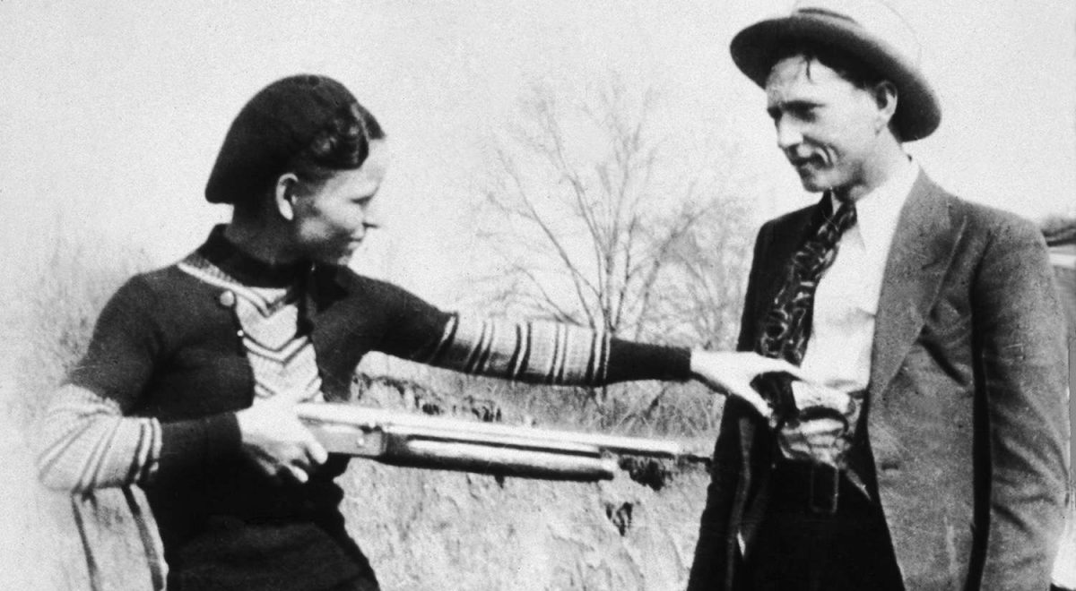 Bonnie Parker jokingly points rifle at Clyde Barrow in black and white historic photo.