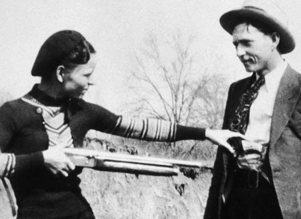 Bonnie Parker jokingly points rifle at Clyde Barrow in black and white historic photo.
