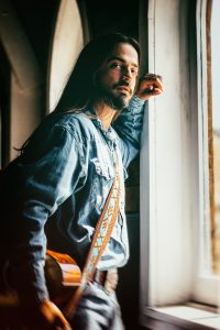 Ian Munsick gazes out a window with his guitar in hand.