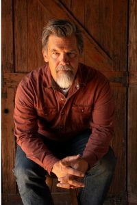 Josh Brolin sits on a stool with jeans and a rust red pearl snap shirt against the backdrop of a barn door.
