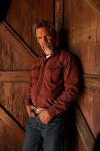 Josh Brolin leans against a barn door, wearing a rust red pearl snap shirt, a silver belt buckle, and jeans.