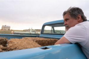 Josh Brolin leans on a bright blue truck loaded with hay bales.