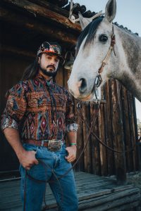 Ian Munsick stands outside of a log cabin next to a horse. He holds the reins of a horse in his left hand and has both thumbs in his pockets.