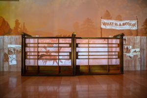 A "Find Your West" art piece featuring a bucking chute gate glowing with projections of historical rodeo images, with a projected banner reading "What is a rodeo?" projected behind the gate.