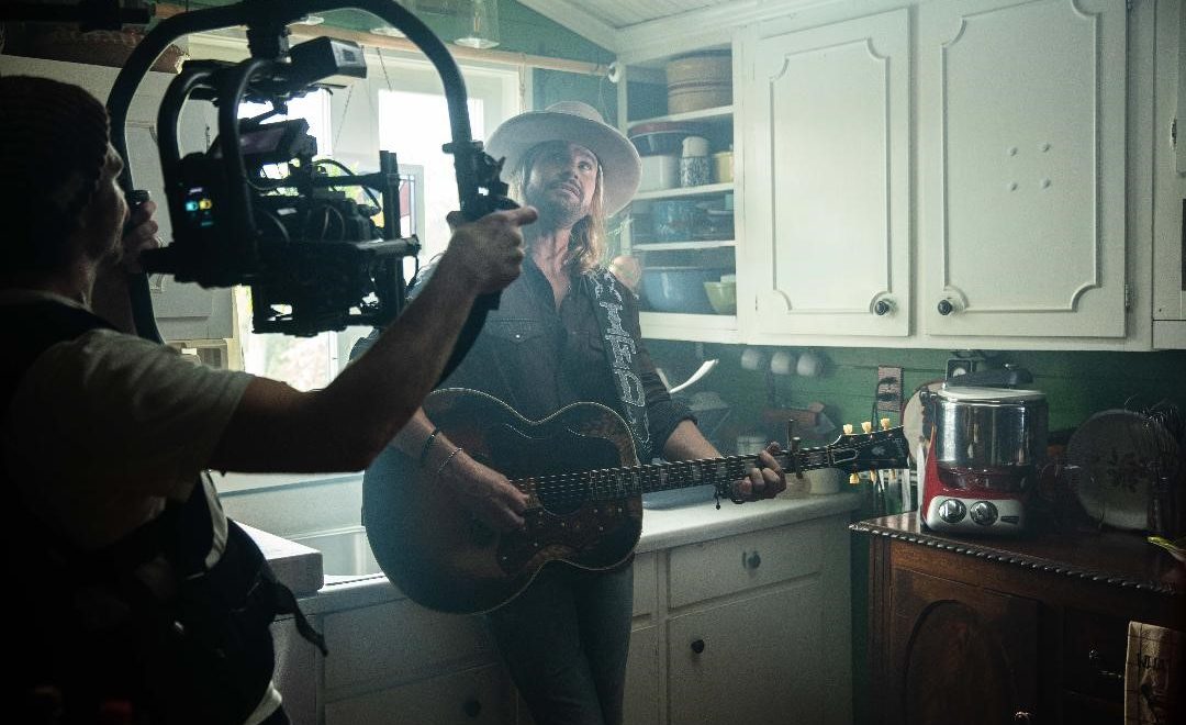 Ira Dean strums his guitar in a kitchen setup while a camera points at him during the filming of a music video.