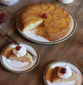 A pineapple upside-down cake on a platter with two slices taken out and placed on individual plates and topped with whipped cream and cherries.