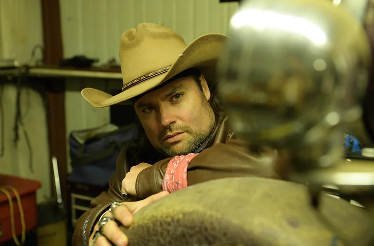Wil Wesley sits with one hand on his guitar case, wearing a hazel cowboy hat, brown leather jacket, multiple silver rings, and a red bandana tied around his wrist.