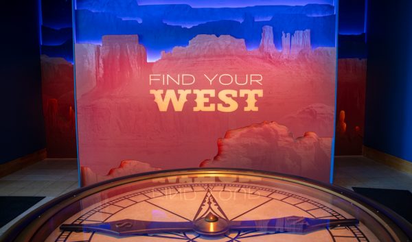 An illuminated sign glows with an artistic interpretation of the Grand Canyon with large yellow letters reading "Find Your West" in the foreground.