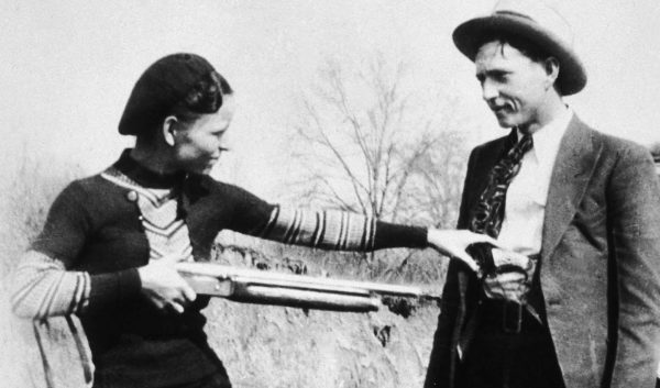 Bonnie Parker jokingly points rifle at Clyde Barrow in black and white historic photo.