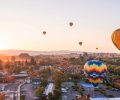 A bird's eye view of a bustling town with shops along the street, homes in rows, hot air balloons taking off into the skies, and a mountain scape in the background as the sun sets.