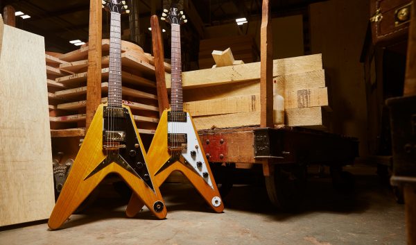 Two Gibson Custom Shop 1958 Korina Flying V Reissues (one in Black and White) behind the backdrop of a Gibson warehouse.
