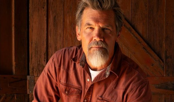 Josh Brolin sits on a stool with jeans and a rust red pearl snap shirt against the backdrop of a barn door.